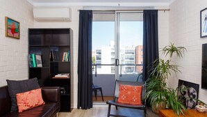 A modern living room with a dark leather sofa, a small wooden table, a bookshelf, and a large window with curtains. - Stay OK Central Fremantle (Fremantle)