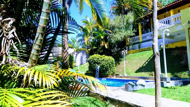 A pool area with a waterfall, surrounded by lush greenery and palm trees. - Country house in Igaratá, standard model, with cable TV, internet, swimming pool, and gourmet area. (IGARATA)