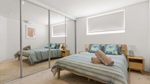 A modern bedroom with a large mirror, a bed with striped bedding, and a nightstand with a lamp. - Salty Steves by Experience Jervis Bay (Huskisson)