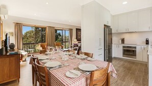 A spacious dining area with a large table set for six, a kitchen with white cabinets, and a balcony with curtains. - Alva Lee by Experience Jervis Bay (Vincentia)