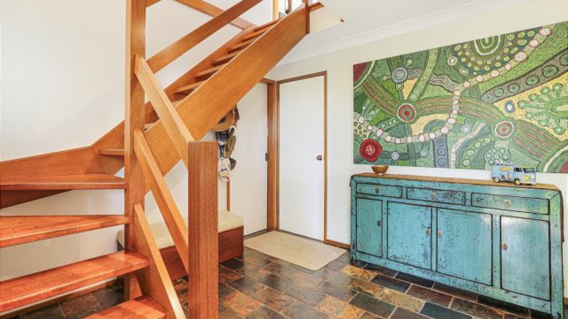 Un escalier en bois menant à une porte, un sol carrelé et un meuble bleu orné d’un tableau décoratif au-dessus.