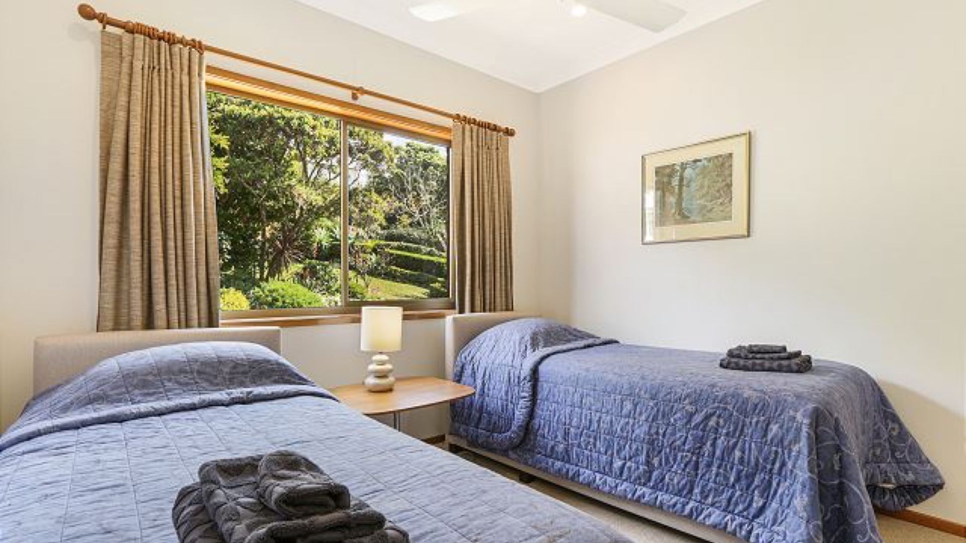 A hotel room with two beds, a window with curtains, a bedside table with a lamp, and a framed picture on the wall.