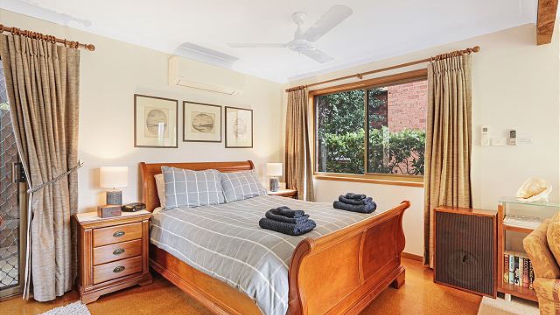 Un dormitorio con una cama de madera, una mesita de noche, un aparador, una ventana con cortinas y un ventilador de techo.
