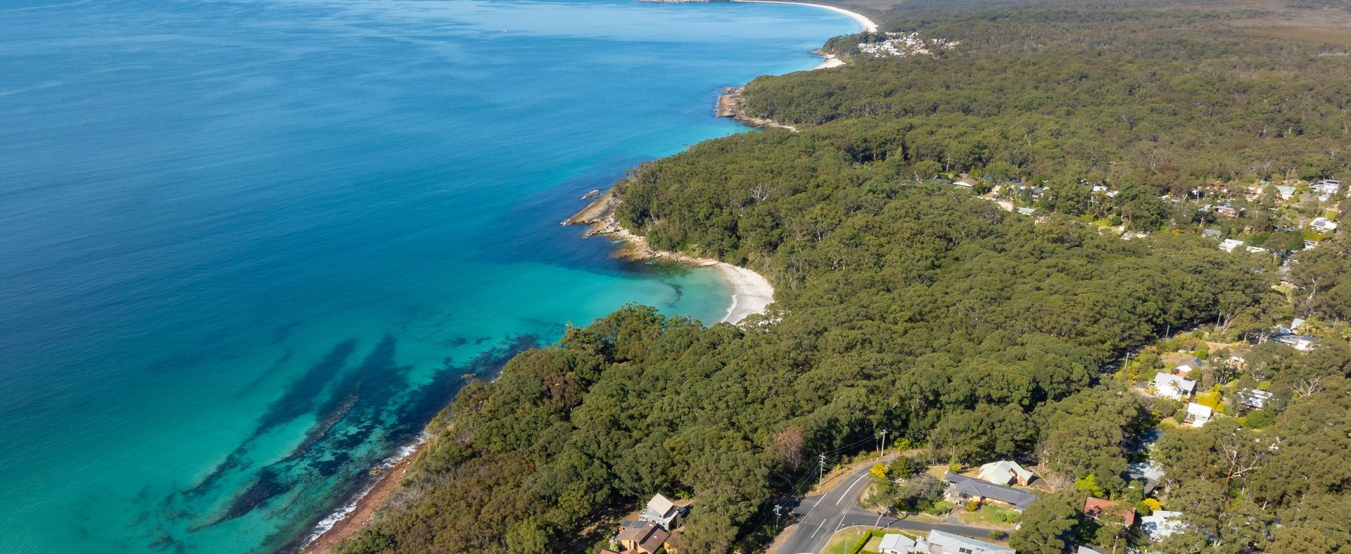 An aerial view of Jervis Bay with a coastal road, residential houses, and a beach.