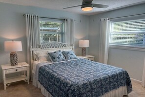 Une chambre à coucher comprenant un lit, deux tables de chevet, un ventilateur de plafond et des fenêtres équipées de stores.