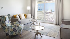 Un salon moderne avec un canapé, un fauteuil à motifs, une petite table ronde avec deux verres, et une vue sur une marina à travers de grandes portes vitrées.