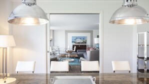 Un salon moderne avec une table basse en verre, des fauteuils blancs et un canapé. On y trouve également un coin repas avec une table et des chaises en verre, ainsi qu’un coin cuisine équipé d’un réfrigérateur et de placards.