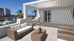 Un balcon agrémenté de meubles en osier, d’un panier de fruits et offrant une vue sur la mer et le paysage urbain.