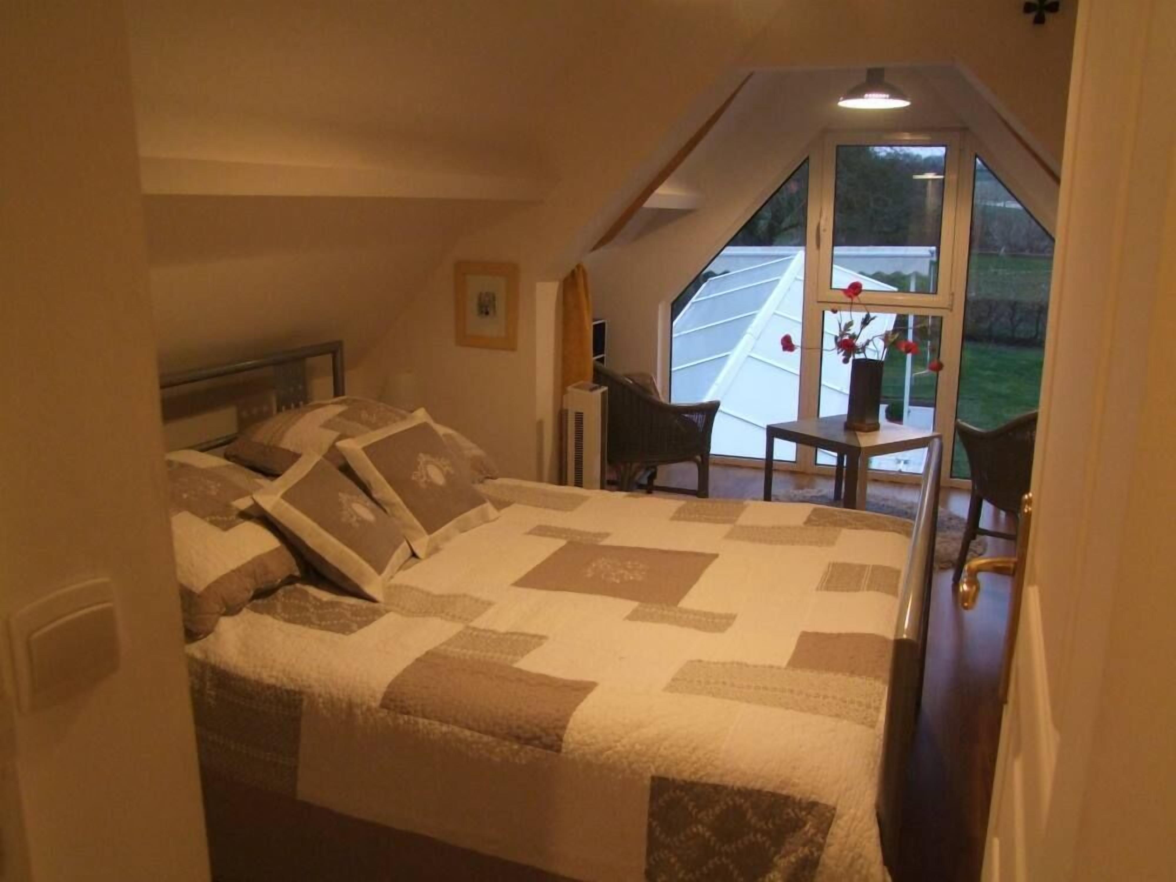 A bedroom with a large bed, a chair, a small table, and a window with a view of a house and greenery.