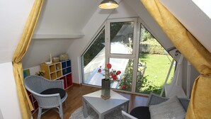 Ein gemütliches Zimmer mit großem Fenster, einem kleinen Tisch, zwei Stühlen und einem Bücherregal.