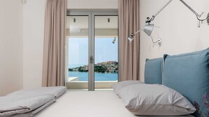 Un dormitorio con cama, dos almohadas, una lámpara de escritorio y vistas a un pueblo costero a través de un ventanal.