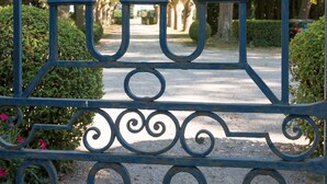 Un cancello in ferro battuto con decorazioni elaborate, che dà accesso a un vialetto alberato e cespugliato.