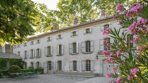Un edificio storico con facciata simmetrica, diverse finestre con persiane e un cortile in ghiaia con sedie in metallo verde.