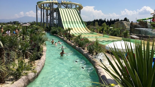 Un parco acquatico con un grande scivolo, piscine cristalline color turchese e una vegetazione rigogliosa.