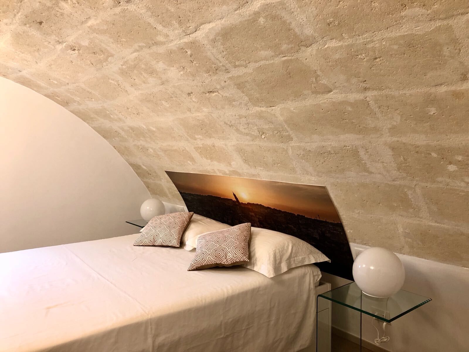 Ein Schlafzimmer mit Steinwand, einem Bett mit gemusterten Kissen, einem Glastisch mit einem weißen, kugelförmigen Objekt und einem gerahmten Bild an der Wand.