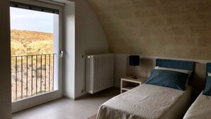 A bedroom with a large window, a radiator, a bedside table with a lamp, and two beds.
