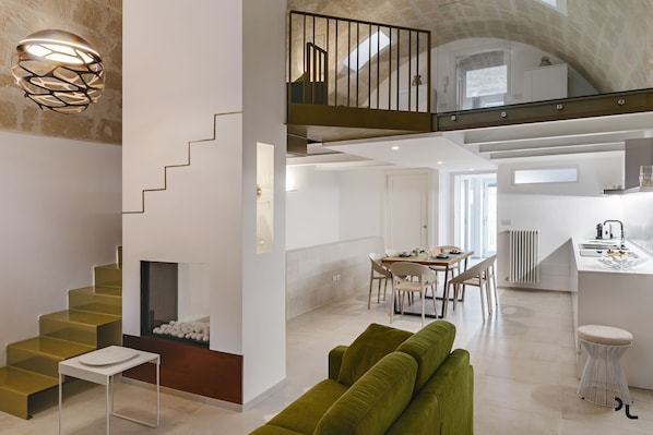 Un interior moderno con escalera, sofá verde, zona de comedor, cocina y un balcón con barandilla.