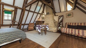 Een loft-achtige kamer met zichtbare houten balken, een stenen open haard, een bank, een eettafel met tafelkleed en een geborduurde spreidek.