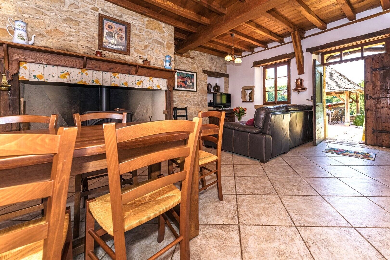Un intérieur rustique avec un mur en pierre, des poutres en bois au plafond, une cheminée, une table à manger avec des chaises, un canapé en cuir et un téléviseur.