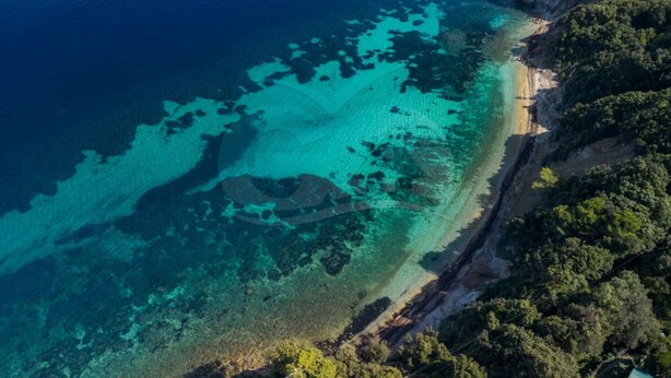 An aerial view of a coastal area with clear turquoise waters, a sandy beach, and dense green vegetation on the cliffs.