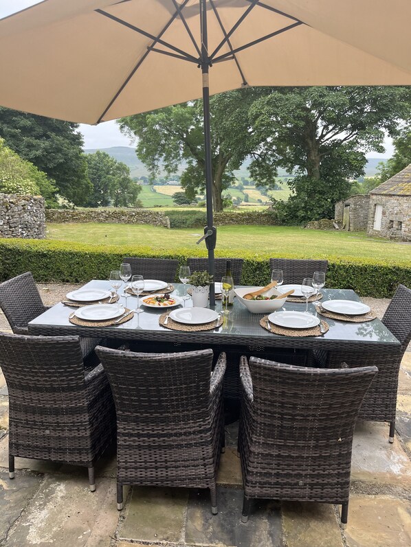 An outdoor dining area with a table set for six, surrounded by wicker chairs, under a large umbrella.