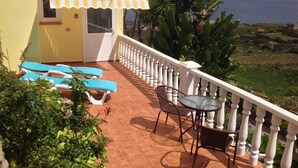 A balcony with blue lounge chairs, a table, and a white railing overlooking a coastal landscape. - AMAZING VIEWS OF ATLANTIC & TEIDE WITH PRIVATE TERRACE , POOL & BBQ (Buen Paso)