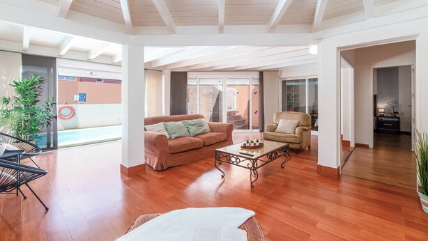 Amplia sala de estar con piso de madera, un sofá marrón, un sillón beige y una mesa de centro. Una ventana grande ofrece luz natural y vistas al exterior.
