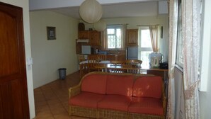 A living room with a wicker sofa, a dining table with chairs, a kitchen area with wooden cabinets, and a window with curtains. - Magnificent sea and mountain views from the roof terrace. Quiet and restful place (Morc. Anna, Flic en Flac)