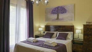 Un lit bien fait, recouvert d’une couvre-lit violette et blanche, avec deux oreillers et des serviettes pliées. On y trouve une table de chevet avec une lampe, une commode en bois et un tableau représentant un arbre accroché au mur.