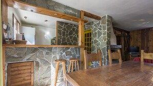 Ein rustikales Interieur mit Steinwand, Holzbalken, einem großen Holztisch und zwei hölzernen Hocker.