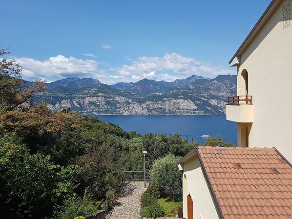 A coastal view with a mountain backdrop, a clear sky, and a pathway leading to a building with a tiled roof.