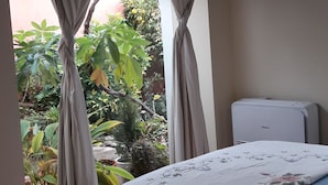 Une chambre à coucher avec un lit, une fenêtre avec des rideaux, un ventilateur de plafond et une vue sur la verdure à l’extérieur.