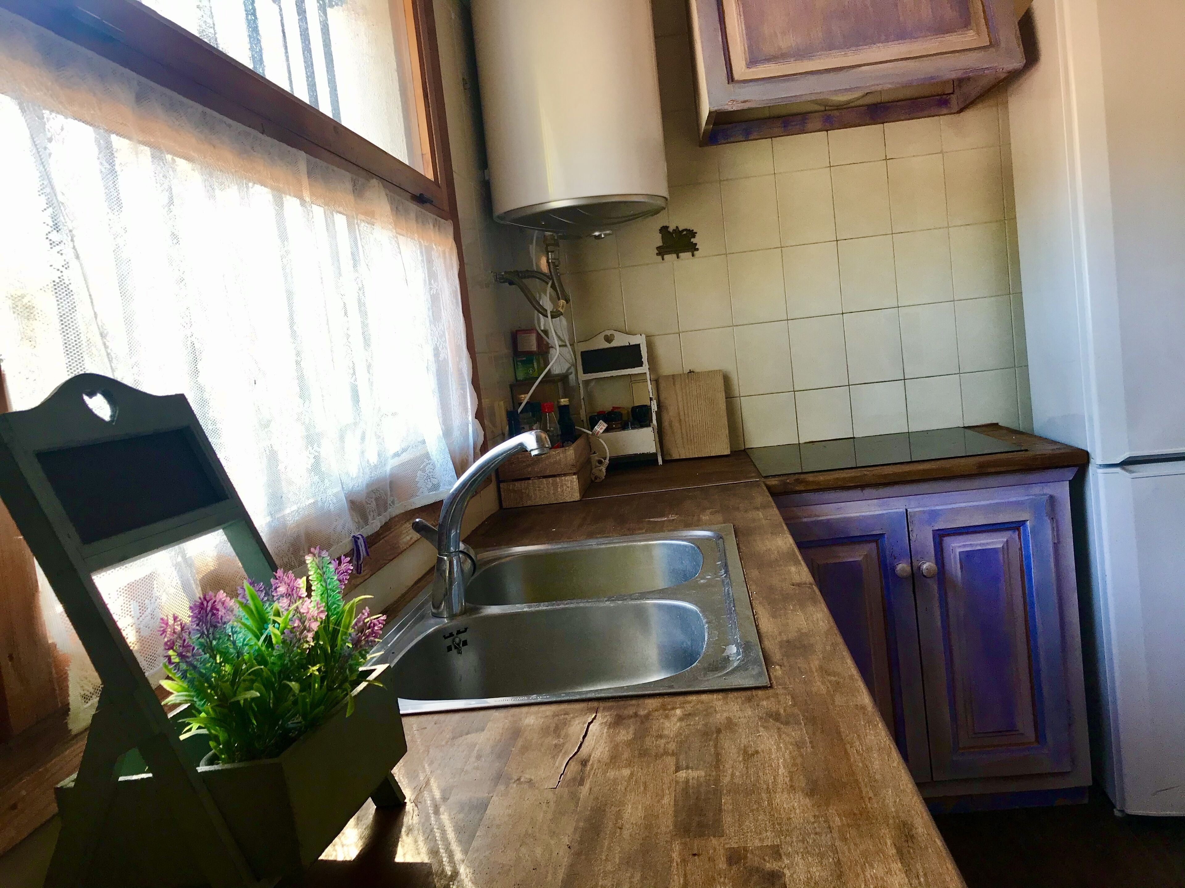Una cocina con encimera de madera, un lavamanos de acero inoxidable y una ventana con cortinas de encaje.