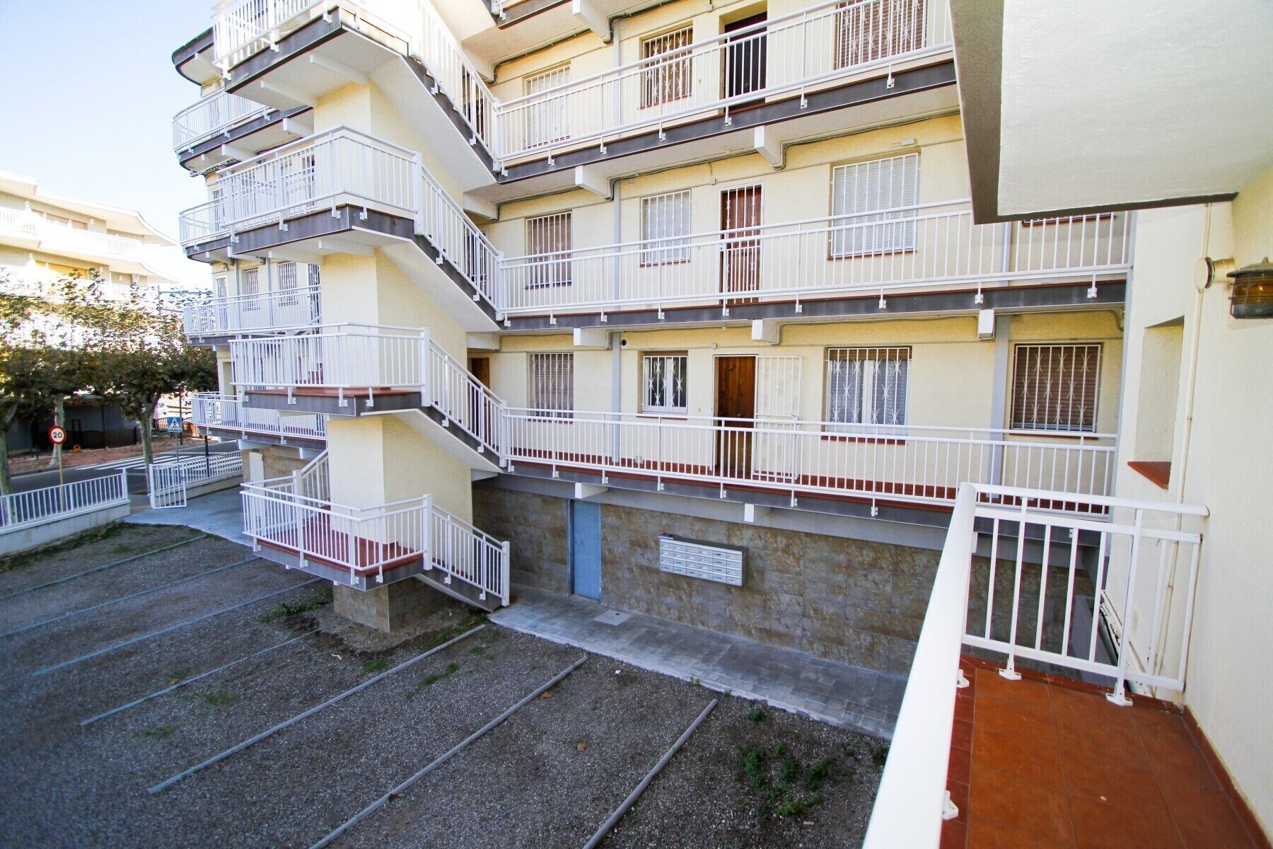 A multi-story residential building with balconies, a gravel driveway, and a red-tiled entrance.