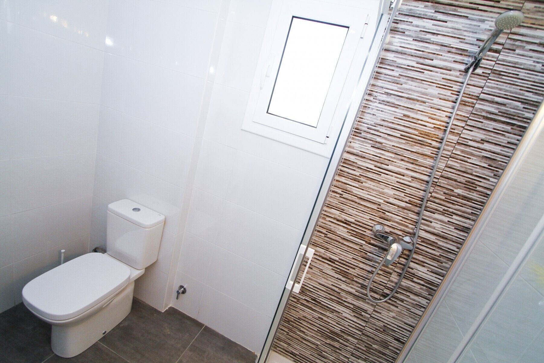 A small, modern bathroom with a toilet, a shower with a wooden panel wall, and a small window.