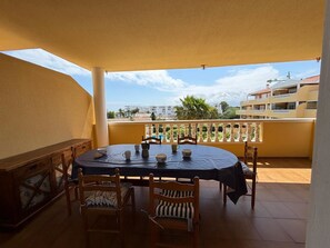 Een overdekt balkon met een ronde tafel en stoelen, met een uitzicht op gebouwen en palmbomen op de achtergrond.