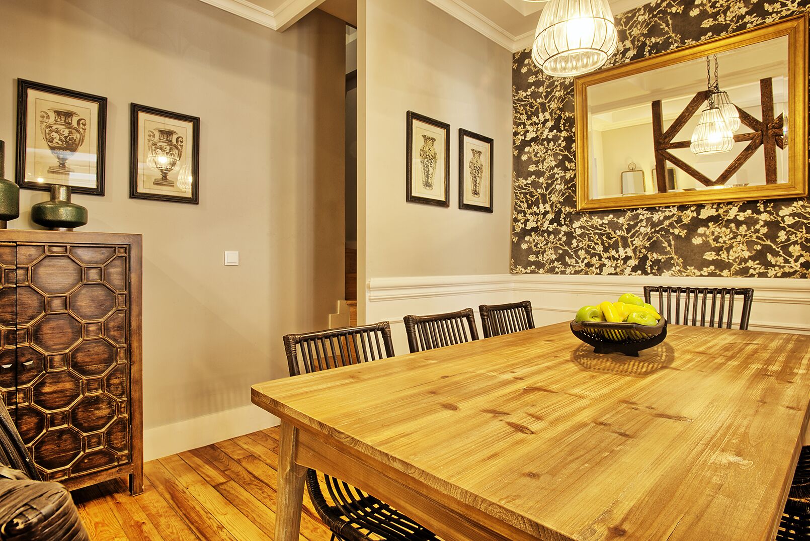 A dining room with a wooden table, chairs, a chandelier, framed artwork, and a decorative wall panel.
