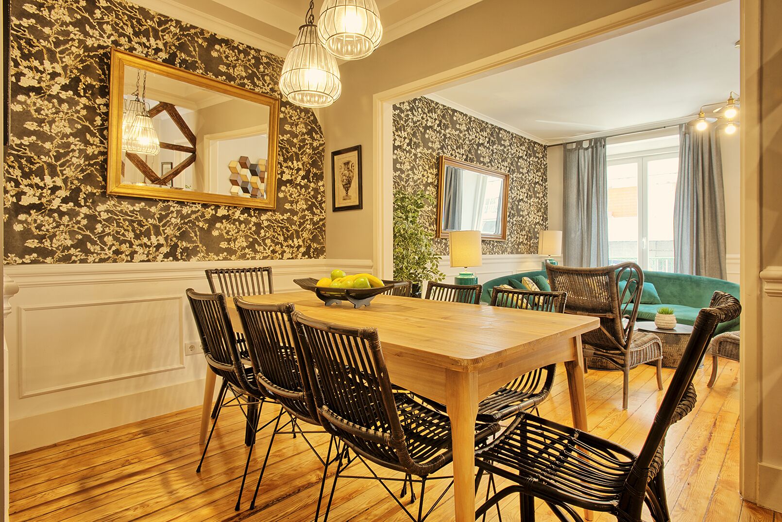 Un comedor con una mesa de madera y sillas de mimbre negro, un sofá verde y un amplio ventanal con cortinas.