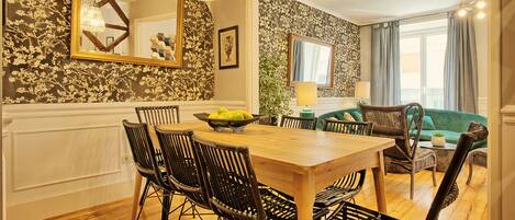 Un comedor con una mesa de madera y sillas de mimbre negro, un sofá verde y un amplio ventanal con cortinas.