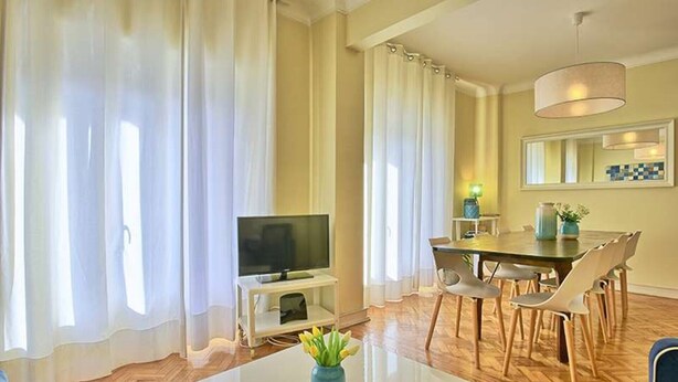 A modern living room with a flat-screen TV, a dining area with a table and chairs, and a large window with sheer curtains.