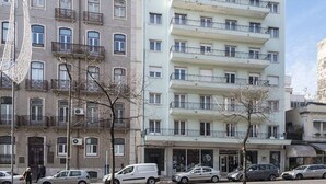 A multi-story residential building with balconies, a ground-level entrance, and parked cars.