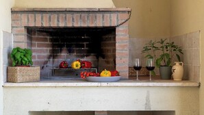 A brick-built outdoor kitchen with a grill, surrounded by stone flooring, baskets, and potted plants. - Podere Collelungo 14, Emma Villas (San Casciano dei Bagni)