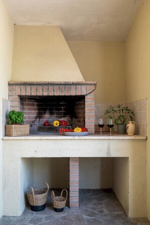 A brick-built outdoor kitchen with a grill, surrounded by stone flooring, baskets, and potted plants.