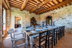 Un espace repas avec une longue table en bois dressée pour le repas, des murs en pierre et un plafond en bois.