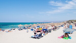 Una playa concurrida con numerosas tumbonas y sombrillas, un cielo azul despejado y un mar tranquilo.