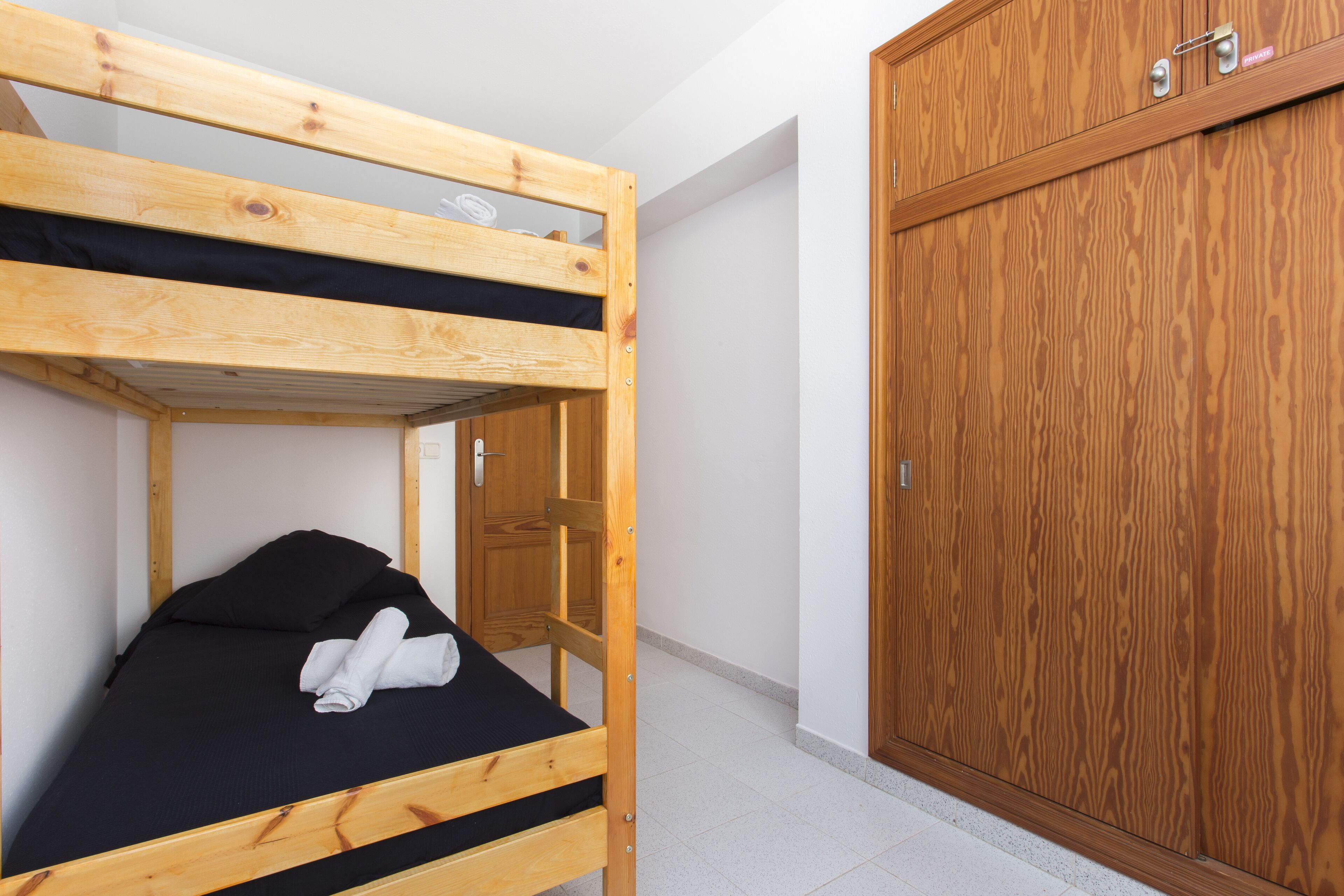Un lit superposé avec un matelas noir et une serviette enroulée, une armoire en bois et une porte blanche dans une pièce aux murs blancs et au sol carrelé.