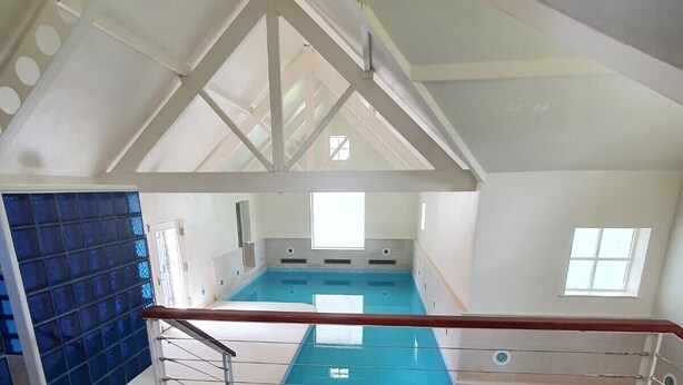 Ein Hallenbad mit Holzdeck im Vordergrund, einer blauen Fliesenwand links und einer hohen Decke mit sichtbaren Balken.