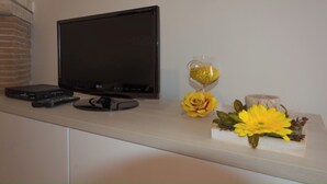 A flat-screen TV, a DVD player, and a decorative hourglass with yellow flowers on a white shelf. - "Between Norba and Norma" cozy apartment (Norma)