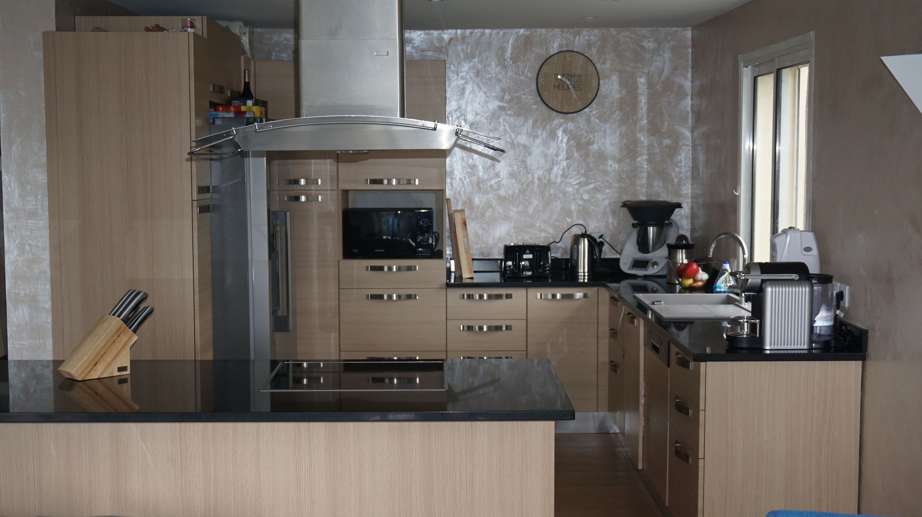 Une cuisine moderne avec des meubles en bois, des appareils électroménagers en acier inoxydable, un four à micro-ondes, une machine à café et une horloge murale.