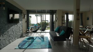Ein modernes Wohnzimmer mit Steinwand, einer Couch mit türkisfarbenen Kissen, einem Esstisch mit Stühlen und Blick auf Palmen durch große Fenster.